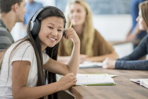 Student smiling, sitting in a classroom environment and listening to audio through headphones