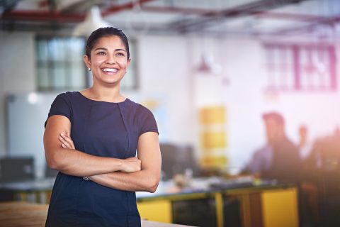 Image of a woman who is smiling and standing in an office environment with her arms crossed